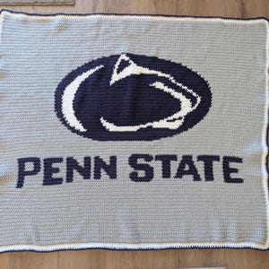 May include: A handmade, crocheted blanket in shades of gray, navy blue, and white. The blanket features the Penn State logo in the center and the words "PENN STATE" across the bottom. The blanket has a white and navy blue border.