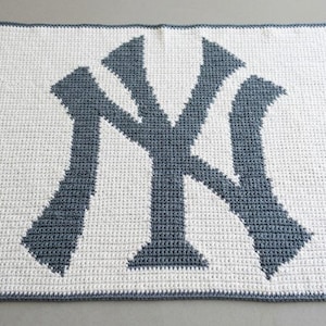 May include: A rectangular crochet blanket featuring a white background with a large, dark grey New York Yankees logo centered in the middle. The blanket is finished with a dark grey border that frames the entire piece, showcasing a consistent stitch pattern throughout the design.