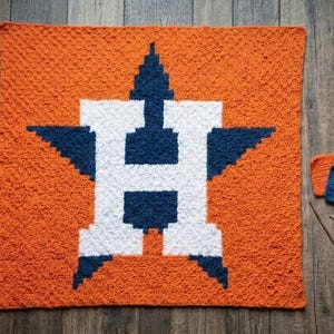 May include: An orange and blue crocheted blanket featuring a large white and blue letter "H" inside a star. The blanket is on a wooden surface, with two small crocheted squares and crochet hooks nearby.