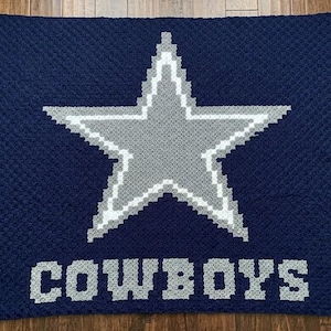 May include: A navy blue crocheted blanket featuring a large gray and white star in the center. The word "COWBOYS" is displayed in white block letters below the star. The blanket is handmade and has a textured appearance.