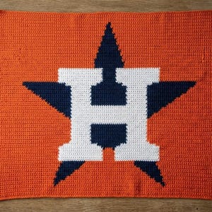 May include: A rectangular crochet blanket featuring a vibrant orange background with a large, centered navy blue star. Overlaid on the star is a bold, white capital letter H, creating a graphic design that spans the center of the textile piece.