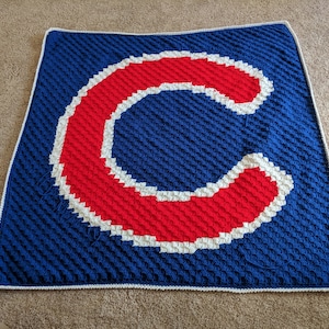 Könnte beinhalten: Eine handgefertigte gehäkelte Decke in Blau, Rot und Weiß. Die Decke zeigt einen großen roten Buchstaben "C" mit weißem Umriss auf blauem Hintergrund. Die Decke hat einen weißen Rand.