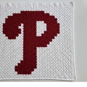 May include: A white and burgundy crocheted blanket featuring a large letter "P" in the center. The blanket has a textured appearance due to the crochet stitches. The "P" is in a solid burgundy color, contrasting with the white background.