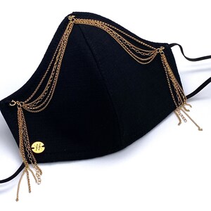 Pu&ograve; includere: Maschera facciale nera con decorazioni a catena dorata. La maschera ha un ciondolo dorato con la lettera "X" su di essa.