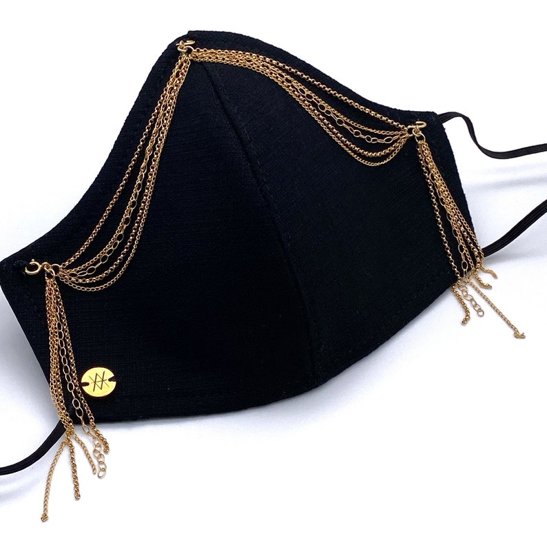 Pu&ograve; includere: Maschera facciale nera con decorazioni a catena dorata. La maschera ha un ciondolo dorato con la lettera "X" su di essa.