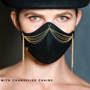 Pu&ograve; includere: Una maschera facciale nera con decorazioni a catena dorata. Le catene pendono dalla maschera, creando un effetto a lampadario. Il testo "WITH CHANDELIER CHAINS" &egrave; stampato sotto la maschera.