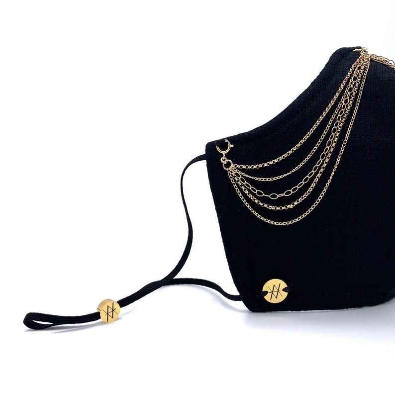 Pu&ograve; includere: Maschera facciale nera con decorazioni a catena dorata e un cinturino elastico nero. La maschera ha un logo circolare dorato con la lettera "H" su ciascuna estremit&agrave; del cinturino.