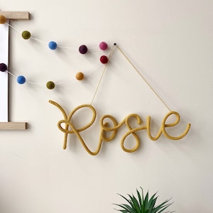 Könnte beinhalten: Ein gelbes, gehäkeltes Namensschild mit der Aufschrift "Rosie", das an einer weißen Wand mit einer Schnur hängt. Das Schild hängt an einem kleinen schwarzen Haken. Links vom Schild hängt eine Girlande aus bunten Filzkugeln an der Wand.