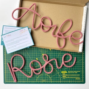 Könnte beinhalten: Zwei rosafarbene, gestrickte Wortzeichen, "Rose" und "Torfe", werden auf einer grünen Schneidematte angezeigt. Die Schilder sind aus dickem Garn gefertigt und haben eine dreidimensionale Optik.