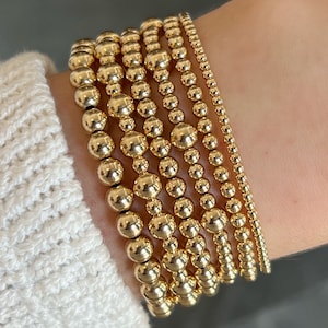 May include: A stack of gold beaded bracelets on a person's wrist. The bracelets are all different sizes and have a shiny finish.