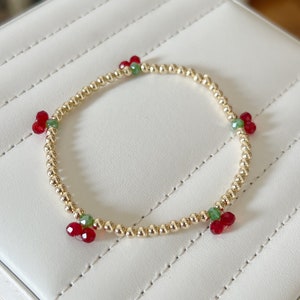 May include: A gold beaded bracelet with red and green crystal accents. The bracelet is on a white surface.