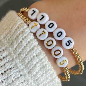 May include: Three gold beaded bracelets with white beads that have black numbers on them. The numbers on the bracelets spell out "17005".