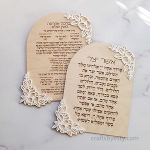 May include: Two wooden plaques with arched tops, featuring Hebrew text and delicate floral cutouts. The plaques are light-colored with dark text, and the floral designs are white. The background is a light marble.