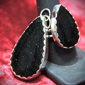 May include: A silver ring featuring two large, teardrop-shaped black stones with sparkling green inclusions. The stones are set in a scalloped silver bezel, creating a unique and eye-catching design. The ring is displayed against a red background.