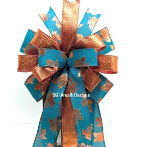 May include: A decorative bow featuring teal and copper-colored ribbons. The teal ribbons are patterned with gold-colored leaves. The bow has multiple loops and streamers, and the text "SG Wreath Designs" is visible.