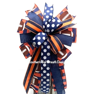 May include: A decorative bow featuring navy blue, orange, and brown ribbons. The bow has a football theme, with football and polka dot patterns. The text "Southern Gal Wreath Design" is visible on the bow.