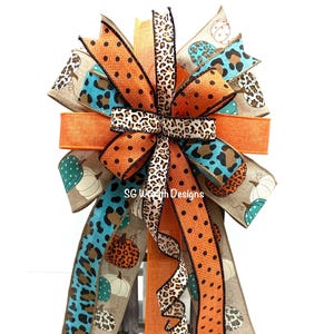 May include: A decorative bow featuring a mix of patterned ribbons in fall colors. The bow includes orange, teal, and leopard print ribbons, with polka dots and pumpkin motifs. The text "SG Wreath Designs" is visible.