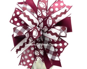 May include: A burgundy and white bow with a football pattern, polka dots, and plaid. The bow is tied around a white lantern. "Southern Gal Wreath Designs" is written on the lantern.