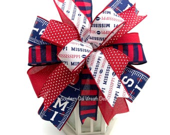 May include: A decorative bow featuring multiple ribbon patterns in red, white, and navy blue. The ribbons include text reading MISSISSIPPI, football graphics, white polka dots on red, navy and red stripes, and markings resembling a football field.