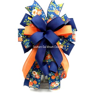 May include: A decorative bow featuring navy blue, orange, and floral patterned ribbons. The bow has a multi-layered design with a large navy blue bow in the center. The text "Southern Gal Wreath Designs" is visible on the bow.