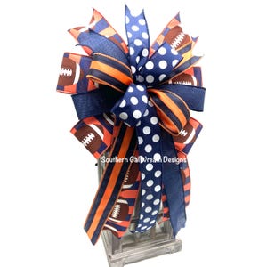 May include: A decorative bow featuring football and polka dot patterns in navy blue, orange, and white. The bow is made of various ribbons and is designed for decorative purposes. The text "Southern Gal Dream Designs" is visible on the bow.