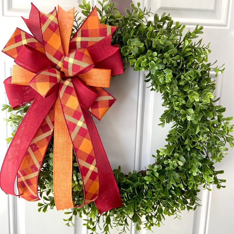 May include: A green boxwood wreath with a large, multi-colored plaid bow. The bow is made of red, orange, and yellow plaid ribbon.