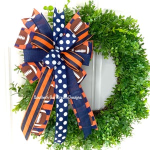 May include: A decorative wreath with a vibrant bow featuring footballs, stripes, and polka dots in navy blue, orange, and white. The wreath is made of lush green foliage, perfect for adding a touch of sporty charm to any home decor.