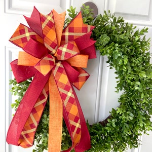 May include: A red and orange plaid bow with a green boxwood wreath. The bow is tied with a large, red ribbon and has a plaid pattern.