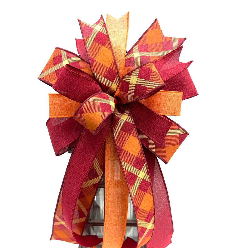 May include: A large, festive bow made with two types of ribbon. One ribbon is a burgundy red with a subtle plaid pattern, and the other is a bright orange with a plaid pattern featuring yellow and orange squares. The bow is tied in a large, full shape with multiple loops.