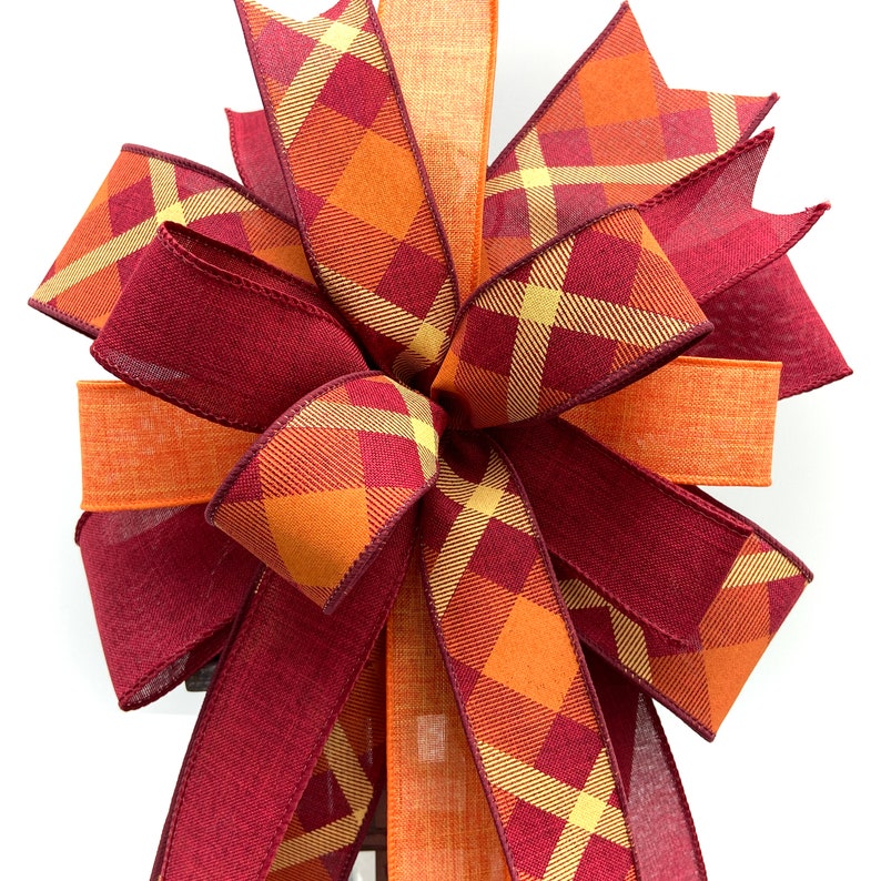 May include: A large, decorative bow made with two types of ribbon. One ribbon is a burgundy color with a subtle sheen, while the other is a plaid pattern with orange, burgundy, and yellow hues. The bow is tied in a classic, symmetrical shape with multiple loops.