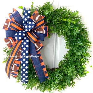May include: A decorative wreath featuring a lush green base and a large bow. The bow is made of navy blue and orange striped ribbon, navy blue polka dot ribbon, and ribbon printed with footballs. The text "Southern Gal Wreath Designs" is visible.