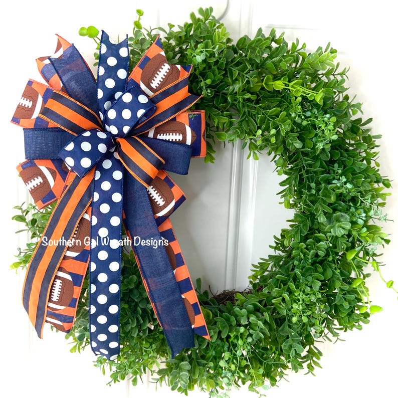 May include: A decorative wreath featuring a lush green base and a large bow. The bow is made of navy blue and orange striped ribbon, navy blue polka dot ribbon, and ribbon printed with footballs. The text "Southern Gal Wreath Designs" is visible.