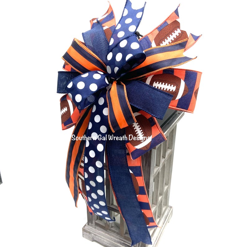 May include: A decorative bow featuring navy blue, orange, and white ribbons. The bow has a football theme, with football graphics and polka dots. The text "Southern Gal Wreath Designs" is visible on the ribbon.