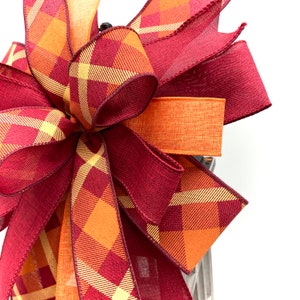May include: A large, festive bow made with burgundy and orange plaid ribbon. The bow is tied with a solid orange ribbon and has a rustic, farmhouse feel.