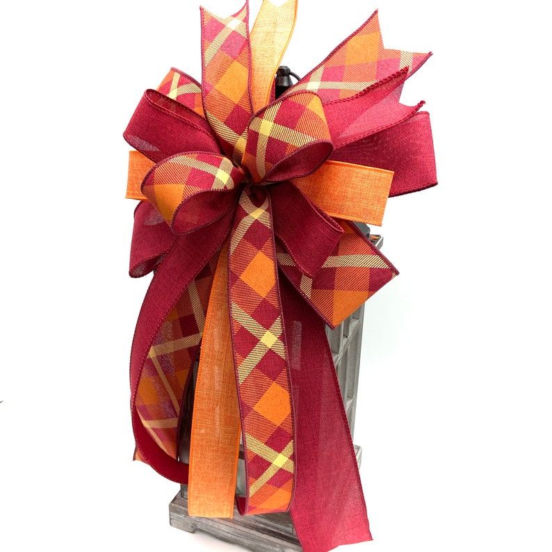 May include: A large, decorative bow made with three different plaid ribbons in shades of burgundy, orange, and yellow. The bow is tied around a lantern.