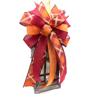 May include: A large, festive bow made with two types of ribbon. The bow is tied with a burgundy ribbon with a plaid pattern in shades of orange, yellow, and burgundy. The bow is tied with a solid orange ribbon. The bow is tied to a wooden lantern.