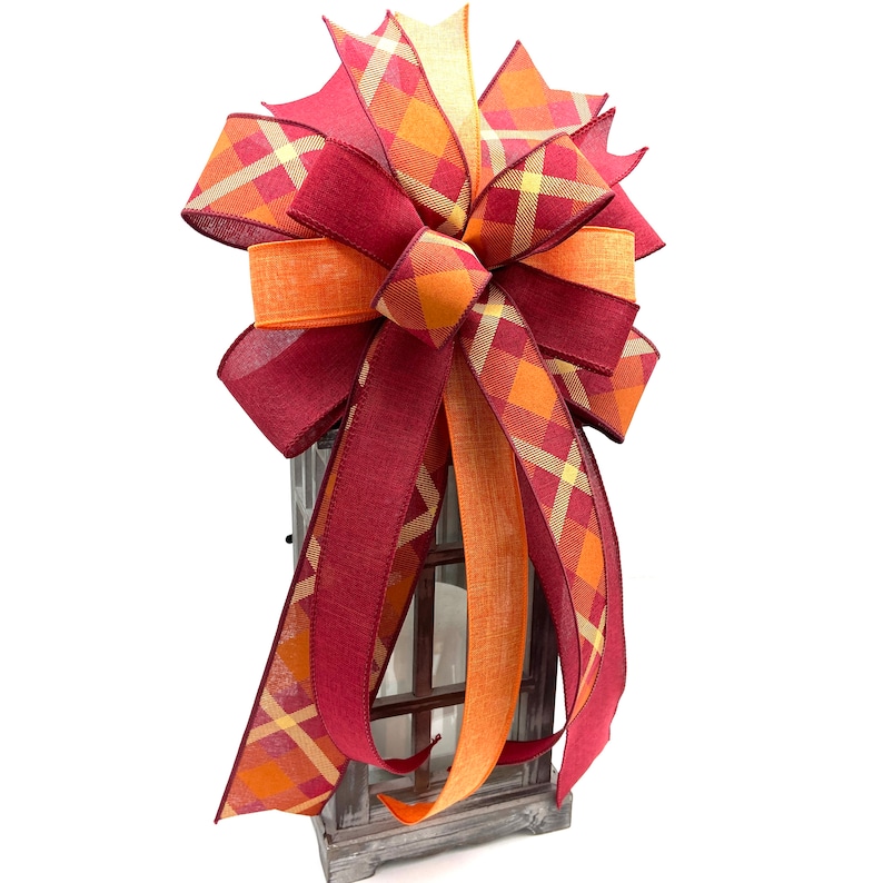 May include: A large, festive bow made with two types of ribbon. The bow is tied with a burgundy ribbon with a plaid pattern in shades of orange, yellow, and burgundy. The bow is tied with a solid orange ribbon. The bow is tied to a wooden lantern.