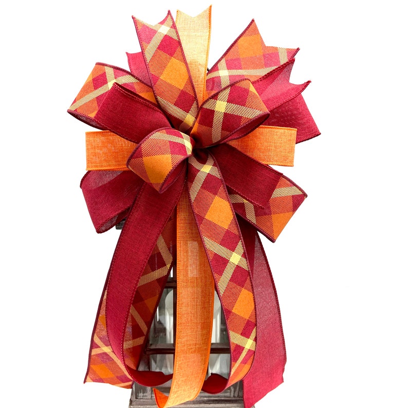 May include: A large, festive bow made with two types of ribbon. One ribbon is a burgundy red with a plaid pattern of orange and yellow. The other ribbon is a solid orange with a subtle texture. The bow is tied in a large, full shape with multiple loops.