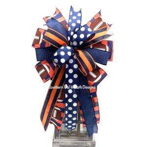 May include: A decorative bow featuring navy blue, orange, and brown ribbons. The bow has a football theme, with football graphics and polka dots. The text "Southern Cal Wreath Designs" is visible.