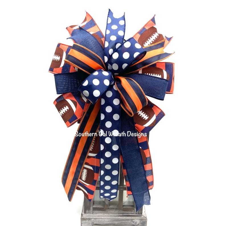 May include: A decorative bow featuring navy blue, orange, and brown ribbons. The bow has a football theme, with football graphics and polka dots. The text "Southern Cal Wreath Designs" is visible.