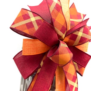 May include: A large bow made with two different plaid ribbons. One ribbon is a burgundy and orange plaid, the other is a yellow and orange plaid. The bow is tied with a loop in the center.