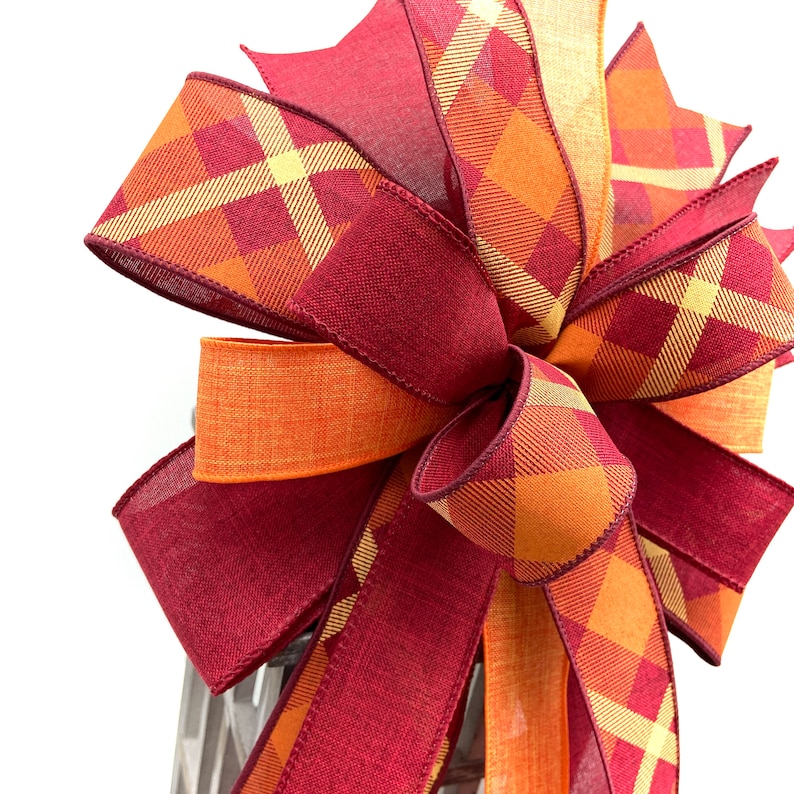 May include: A large bow made with two different plaid ribbons. One ribbon is a burgundy and orange plaid, the other is a yellow and orange plaid. The bow is tied with a loop in the center.
