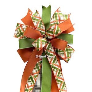 May include: A decorative bow featuring a mix of solid and patterned ribbons. The bow incorporates shades of orange, green, and a plaid pattern with white, orange, and green. The bow is likely for seasonal or holiday decor.