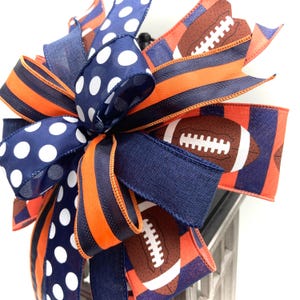 May include: A decorative bow featuring a football theme. The bow incorporates navy blue, orange, and white polka dot ribbons. Some ribbons display footballs. The bow is suitable for various decorative purposes.