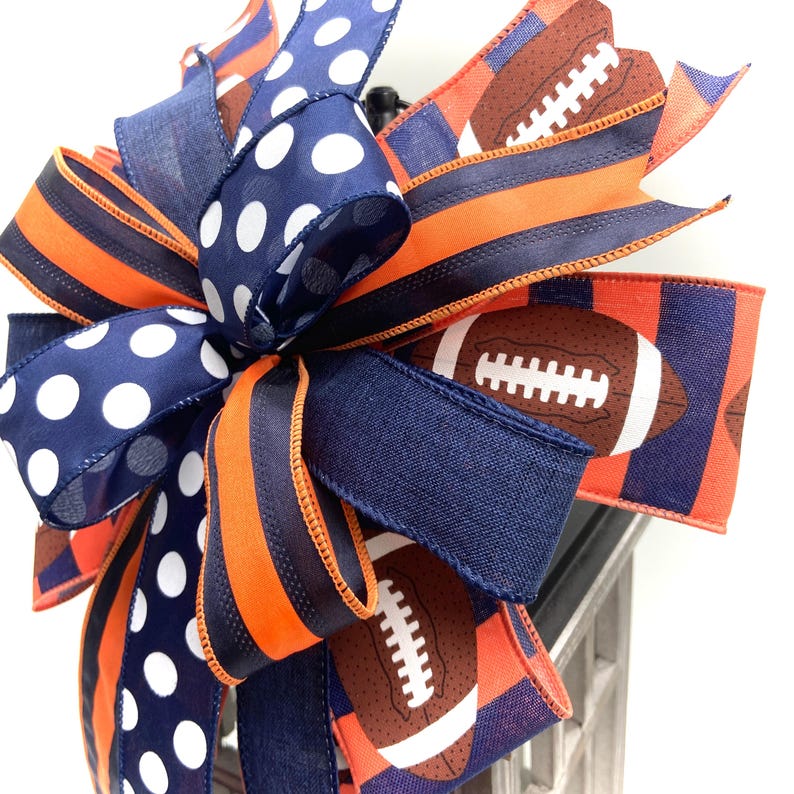 May include: A decorative bow featuring a football theme. The bow incorporates navy blue, orange, and white polka dot ribbons. Some ribbons display footballs. The bow is suitable for various decorative purposes.