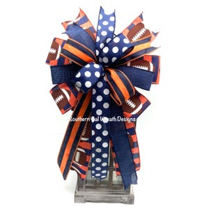 May include: A decorative bow featuring footballs, stripes, and polka dots in navy blue, orange, and white. The bow is made of various ribbons and is designed for decorative purposes. The text "Southern Gal Wreath Designs" is visible on the bow.