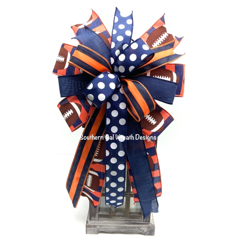 May include: A decorative bow featuring footballs, stripes, and polka dots in navy blue, orange, and white. The bow is made of various ribbons and is designed for decorative purposes. The text "Southern Gal Wreath Designs" is visible on the bow.