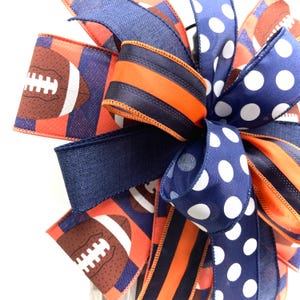 May include: A decorative bow featuring a football theme. The bow incorporates ribbons in navy blue, orange, and white. Some ribbons display footballs, while others have polka dots or stripes. The bow is suitable for sports-themed decorations.