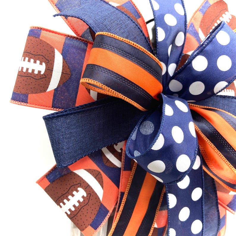 May include: A decorative bow featuring a football theme. The bow incorporates ribbons in navy blue, orange, and white. Some ribbons display footballs, while others have polka dots or stripes. The bow is suitable for sports-themed decorations.
