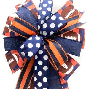 May include: A decorative bow featuring a football theme. The bow has navy blue, orange, and white polka dot ribbons. Some ribbons display footballs. The bow is suitable for various crafts and decorations.
